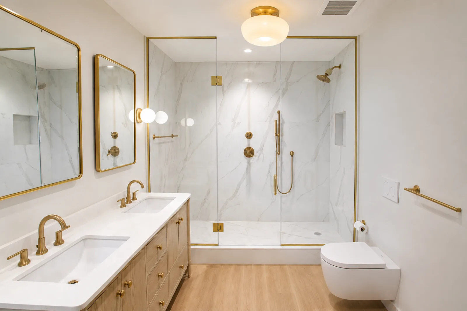 A warm white NYC bathroom with brushed gold fixtures, a marble-look walk-in shower, a light oak double vanity, and a wall-hung toilet