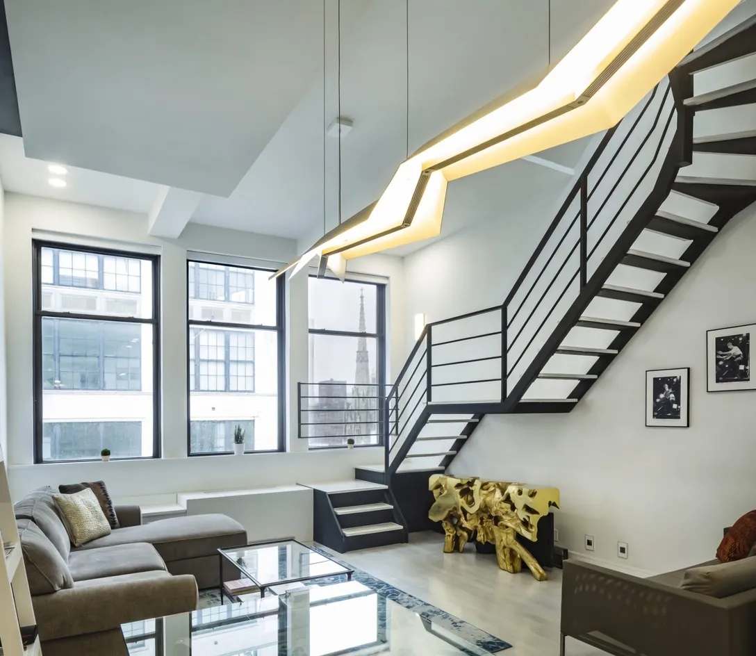 A dramatic double-height NYC loft living room with a sculptural floating staircase and industrial windows