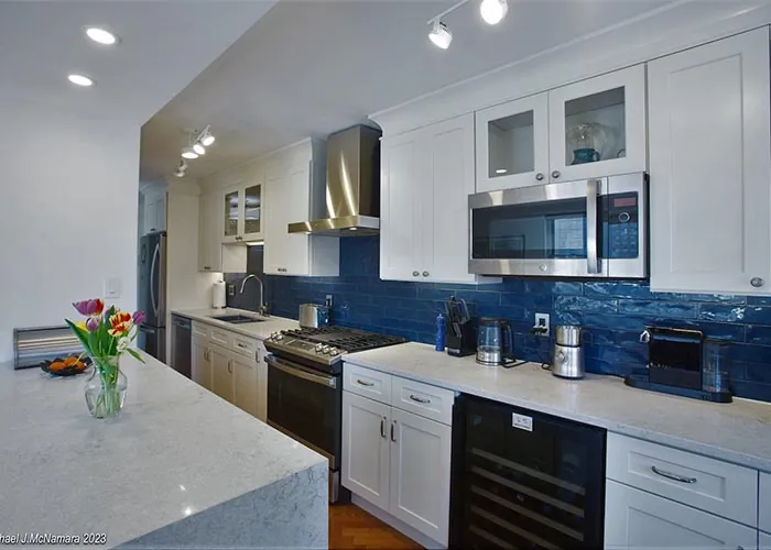 A renovated white shaker kitchen with gray-veined quartz counters, a glossy blue subway tile backsplash, and a wine refrigerator